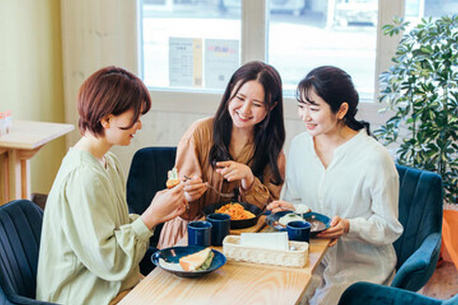 ビジネスウーマン3人が洋食ランチを談笑しながら食べている。