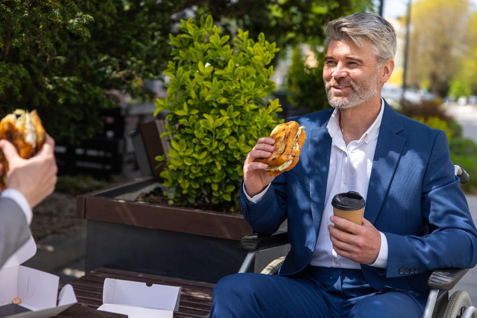 A man sits outside on a bench with a colleague having a hamburger lunch.