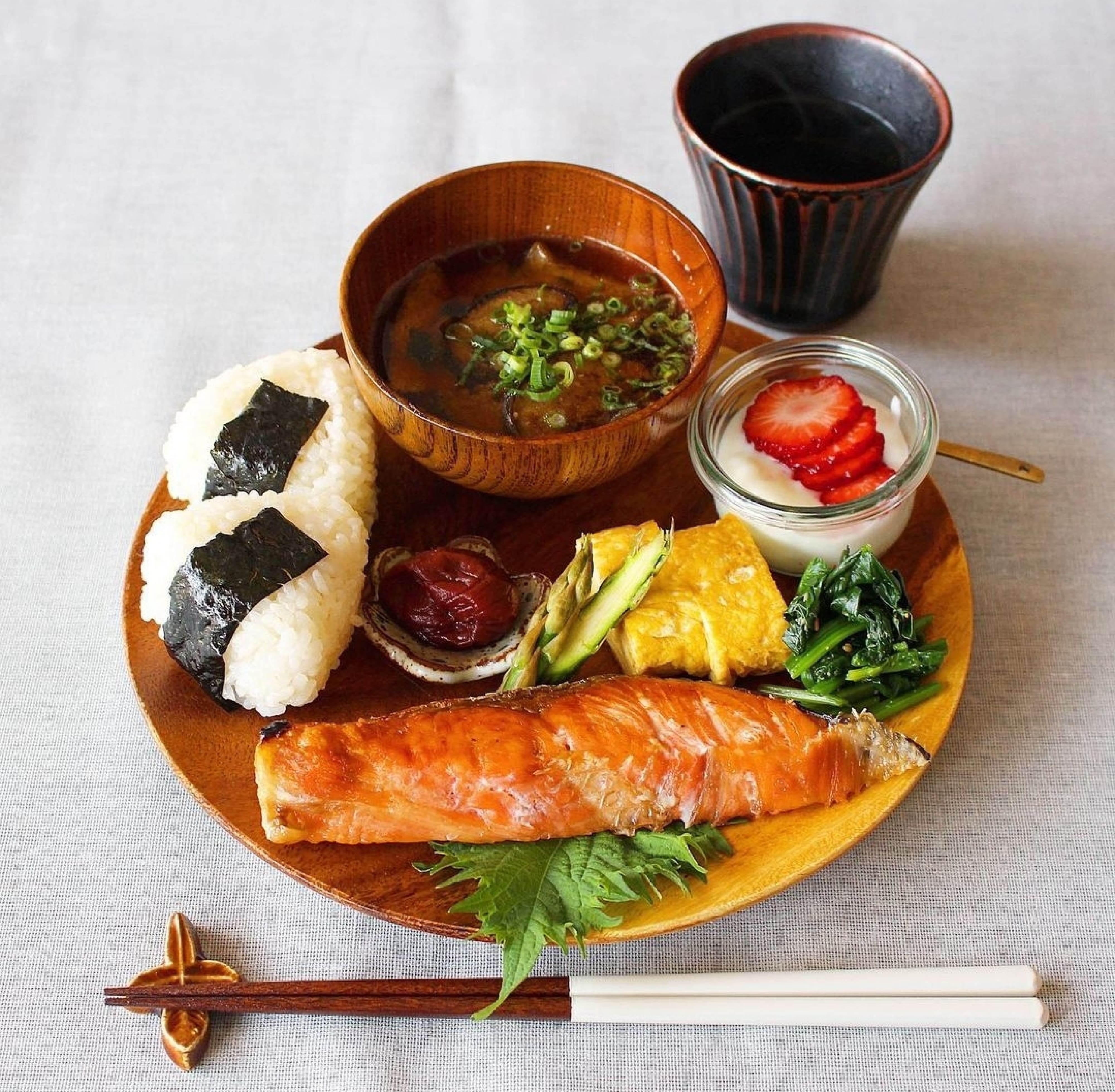 味噌汁、焼き鮭、卵焼き、おにぎり、いちごをのせたヨーグルトがプレートにのっている日本の朝食。