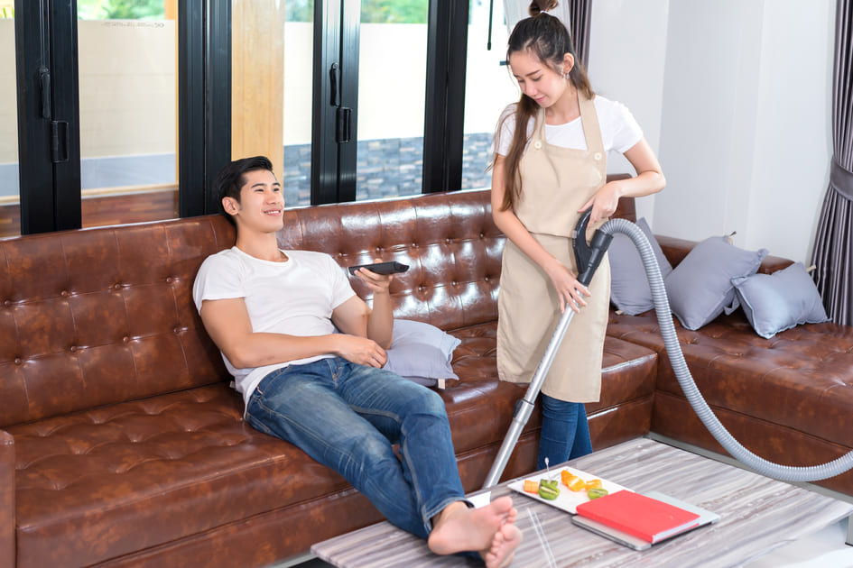 夫がソファーに座りテレビを楽しんでいるそばで掃除機をかける妻。